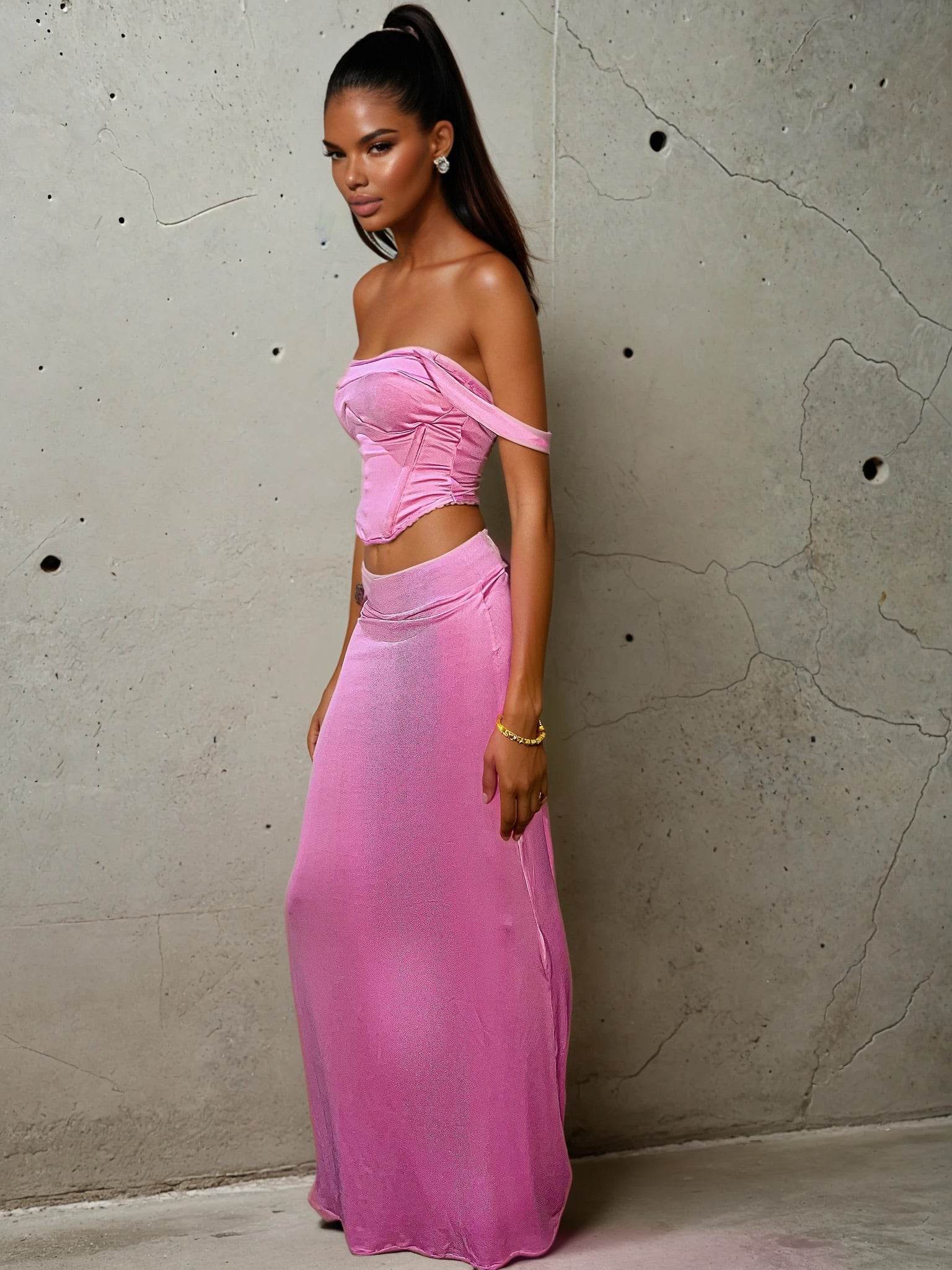 Model wearing a pastel pink two-piece set featuring a structured corset-style crop top with wide straps and visible boning, paired with a form-hugging maxi skirt. The satin-like fabric gives a soft shimmer under light, accentuating the romantic yet modern silhouette. Styled with loose waves and a neutral makeup look, set against an urban concrete wall.