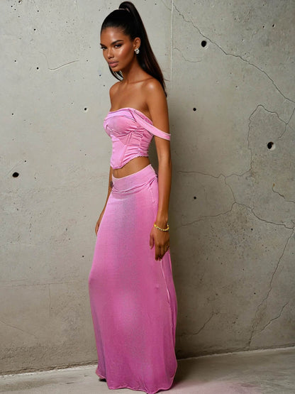 Model wearing a pastel pink two-piece set featuring a structured corset-style crop top with wide straps and visible boning, paired with a form-hugging maxi skirt. The satin-like fabric gives a soft shimmer under light, accentuating the romantic yet modern silhouette. Styled with loose waves and a neutral makeup look, set against an urban concrete wall.