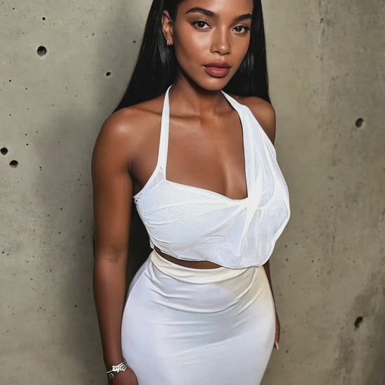 Model wearing a sleek white halter crop top paired with a high-waisted, form-fitting maxi skirt. The outfit features draped mesh detailing across the bust and a smooth satin-like finish on the skirt, creating a refined and modern silhouette. Styled with soft glam makeup, minimalist jewelry, and straight flowing hair, set against a raw concrete backdrop.
