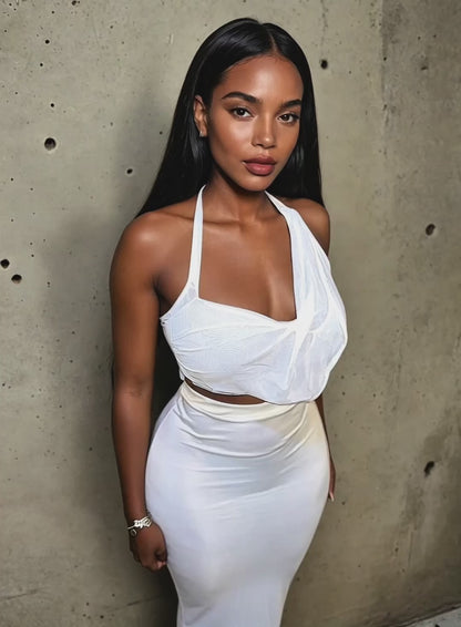 Model wearing a sleek white halter crop top paired with a high-waisted, form-fitting maxi skirt. The outfit features draped mesh detailing across the bust and a smooth satin-like finish on the skirt, creating a refined and modern silhouette. Styled with soft glam makeup, minimalist jewelry, and straight flowing hair, set against a raw concrete backdrop.